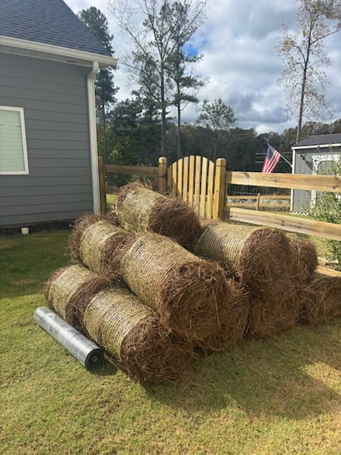 Round bales of pine straw stacked on a lawn beside a new wooden fence, ready to spread